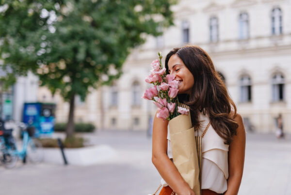 Cheerful young woman holding a bouquet of pink Lisianthus flowers while smiling with closed eyes in a city street, enjoying the scent and the urban landscape
